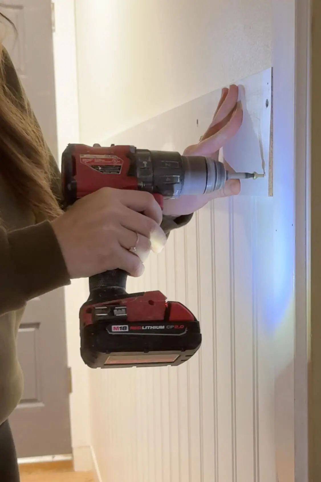  close-up of a woman securing a wooden peg rail onto beadboard paneling with a red cordless drill. Her hand holds the rail in place, highlighting precision and care during the installation process.